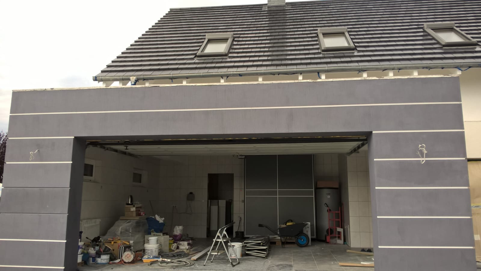 Szara elewacja garażu w trakcie wykończenia, widoczne białe pasy dekoracyjne, narzędzia i materiały budowlane wewnątrz, dach z oknami połaciowymi.