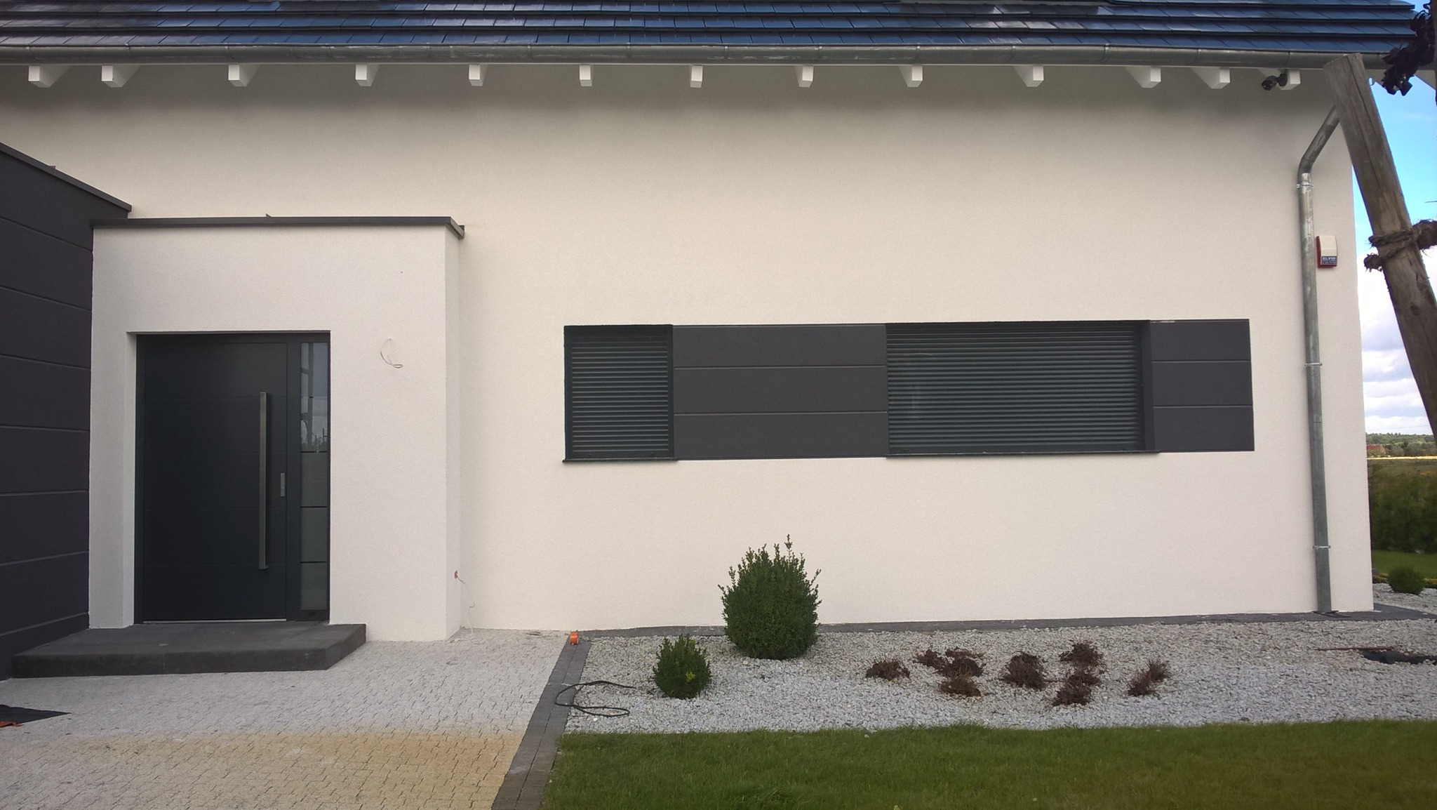 Nowoczesny dom jednorodzinny z jasną elewacją, antracytowymi detalami okiennymi i drzwiami, otoczony ozdobnym żwirem i niską roślinnością.