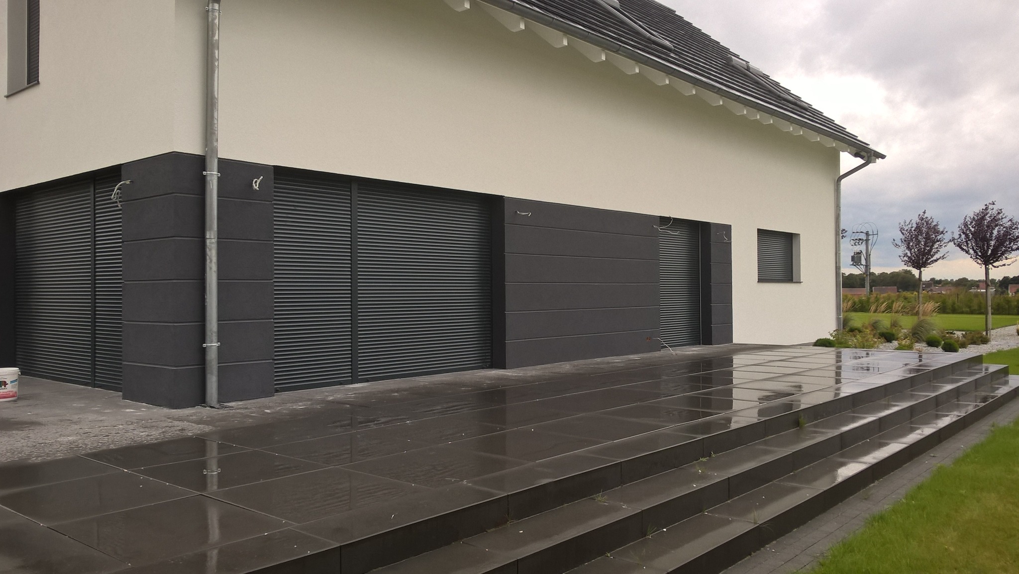 Nowoczesny dom jednorodzinny z elewacją w odcieniach bieli i szarości, z widocznymi żaluzjami zewnętrznymi i schodami wejściowymi wyłożonymi błyszczącymi płytami, po deszczu.