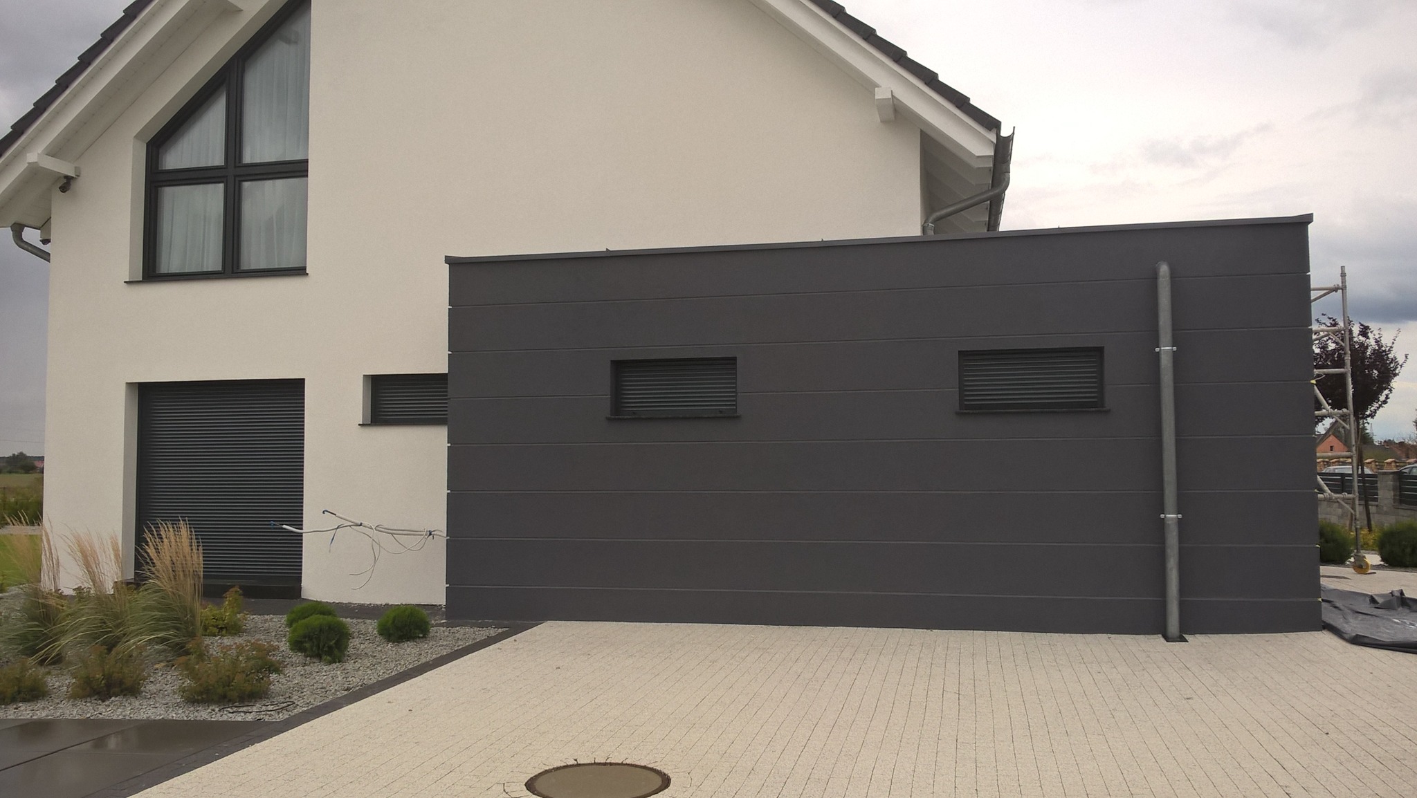 Nowoczesny dom z szarą elewacją panelową i białym tynkiem, widoczne okna z roletami, kostka brukowa na podjeździe i rusztowanie z boku budynku.