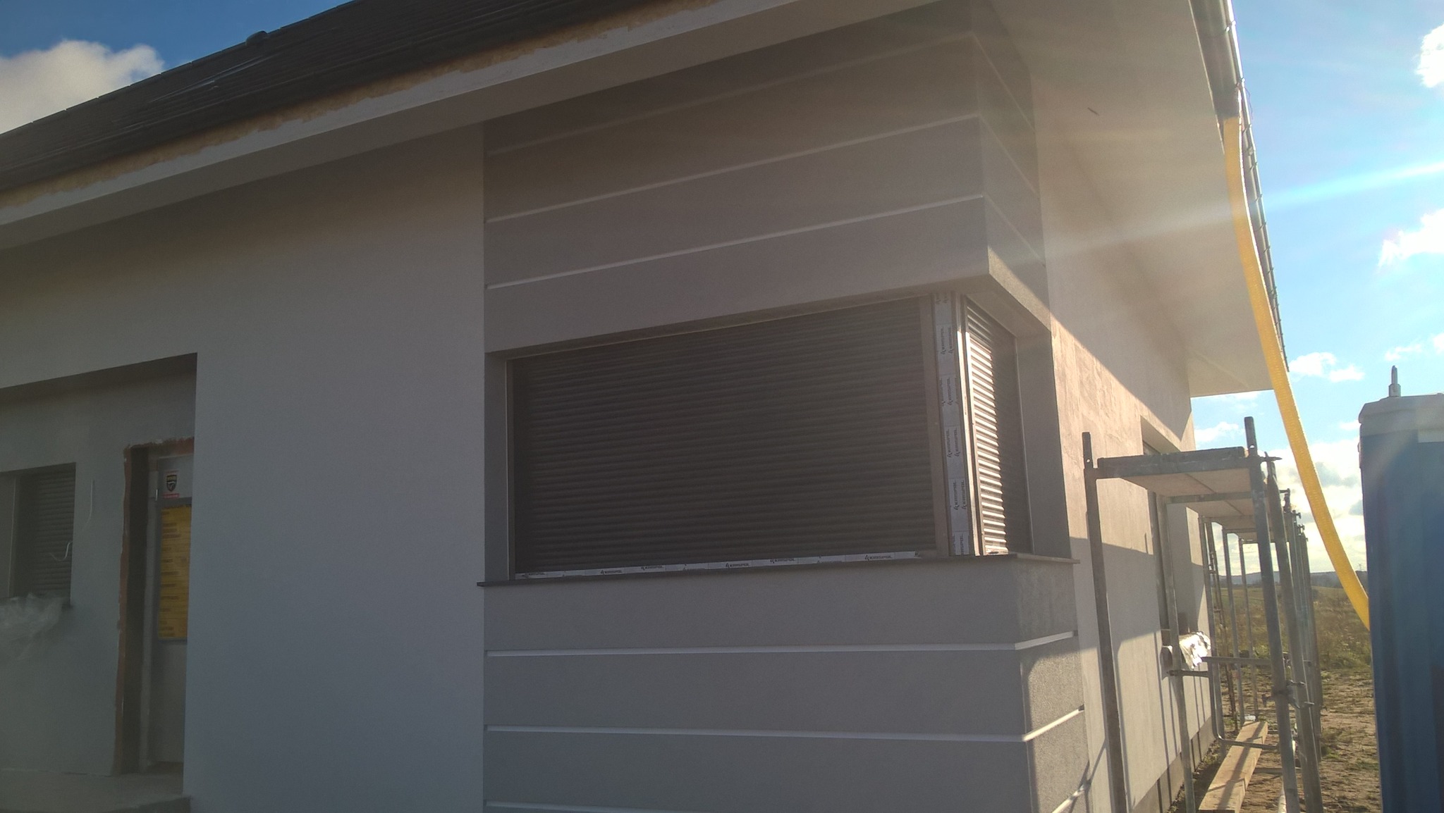 Szara elewacja domu jednorodzinnego z poziomymi frezami, okno z roletą zewnętrzną, fragment rusztowania w słoneczny dzień.