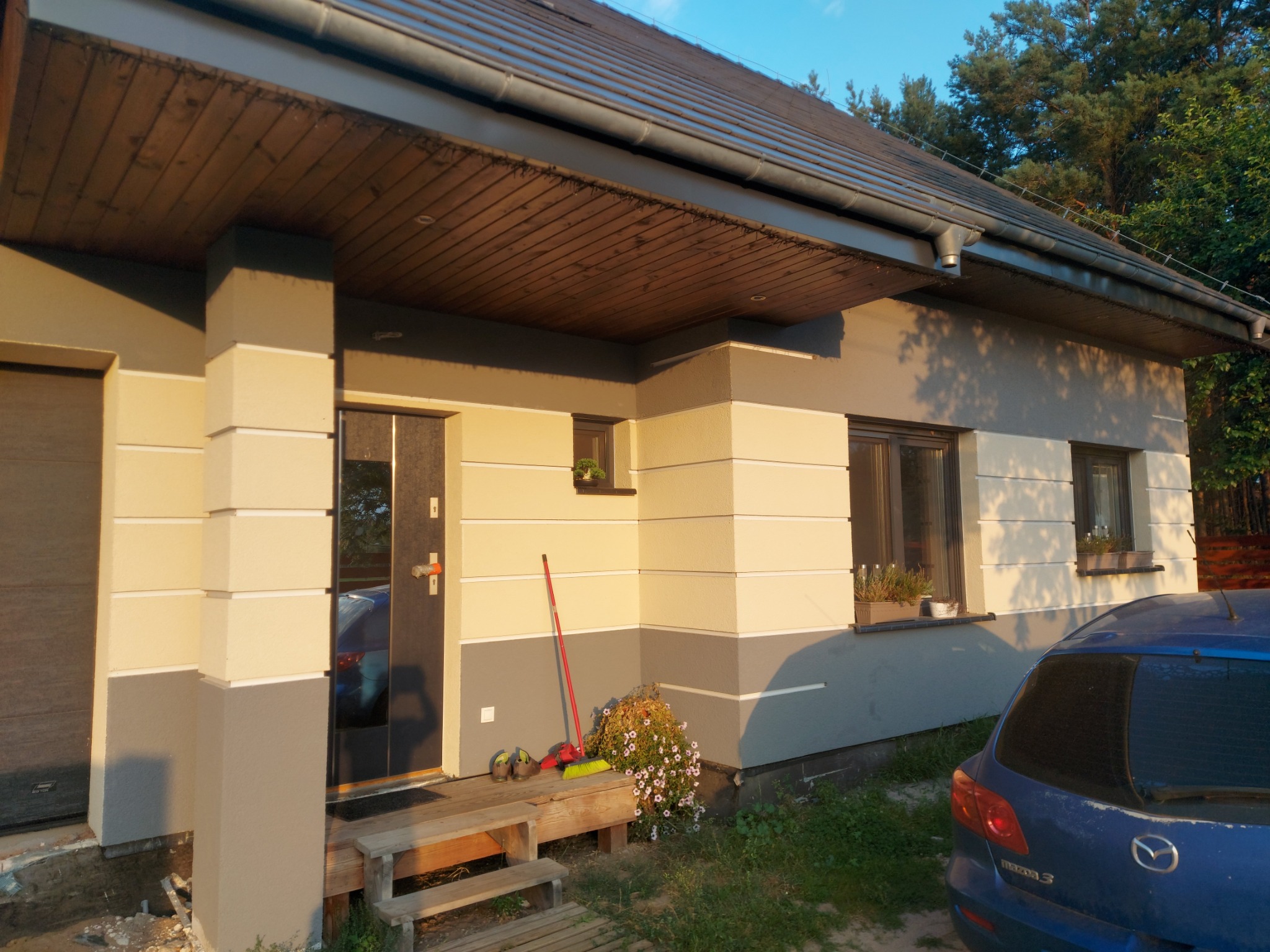 Dwukolorowa elewacja domu jednorodzinnego z widoczną częścią dachu, drewnianym podbitką i schodami wejściowymi; obok wejścia stoi czerwona zmiotka i buty robocze.