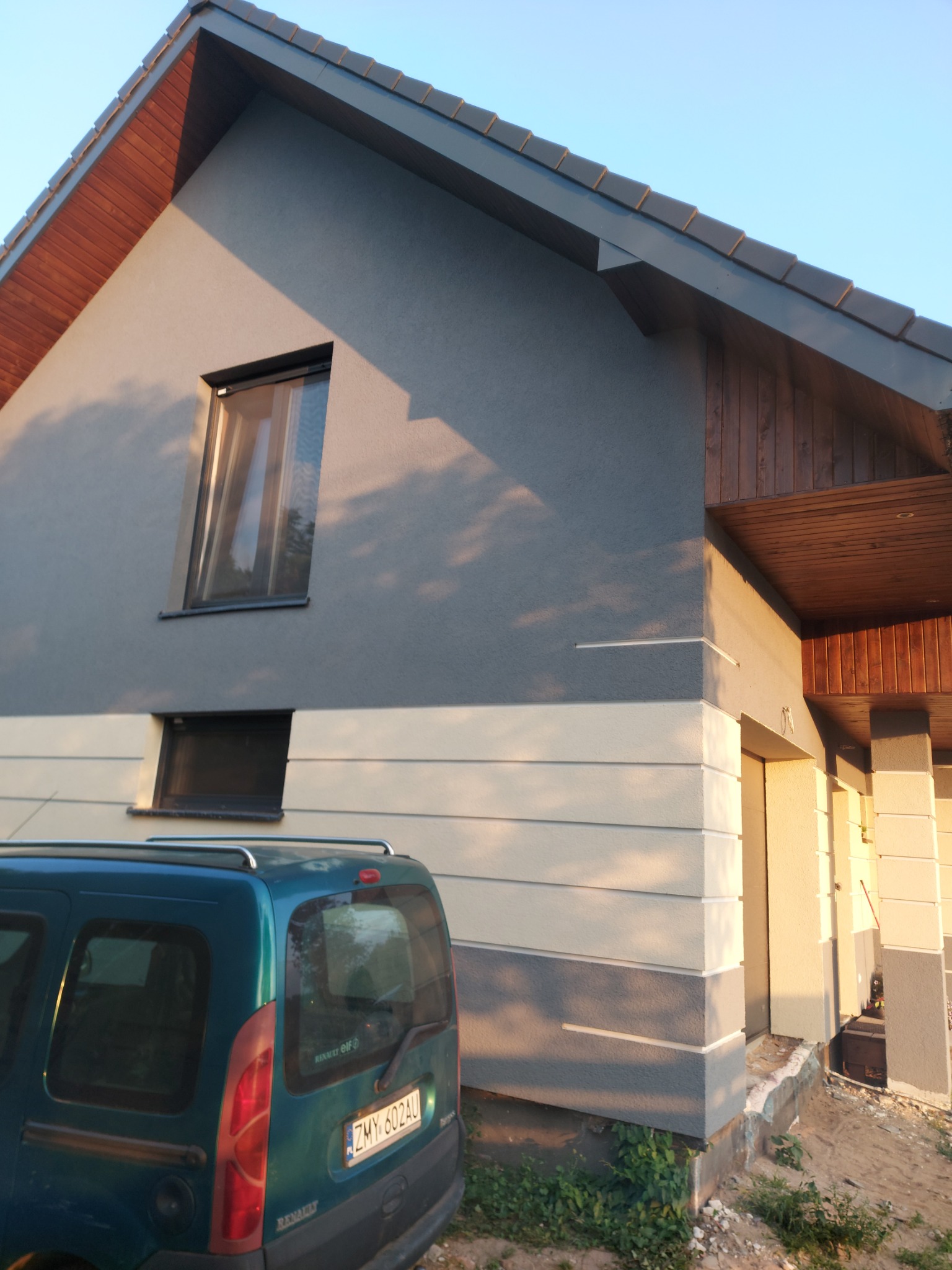 Dwukolorowa elewacja domu z widocznym podziałem na szary tynk na górze i pasy jasnego tynku na dole, z fragmentem zaparkowanego zielonego samochodu Renault Kangoo w kadrze.
