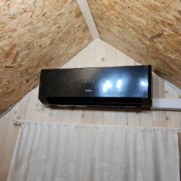 Czarny klimatyzator GREE zamontowany na drewnianej ścianie pod skośnym sufitem z płyty OSB, widoczna biała rura instalacyjna i firanka w kwiaty.