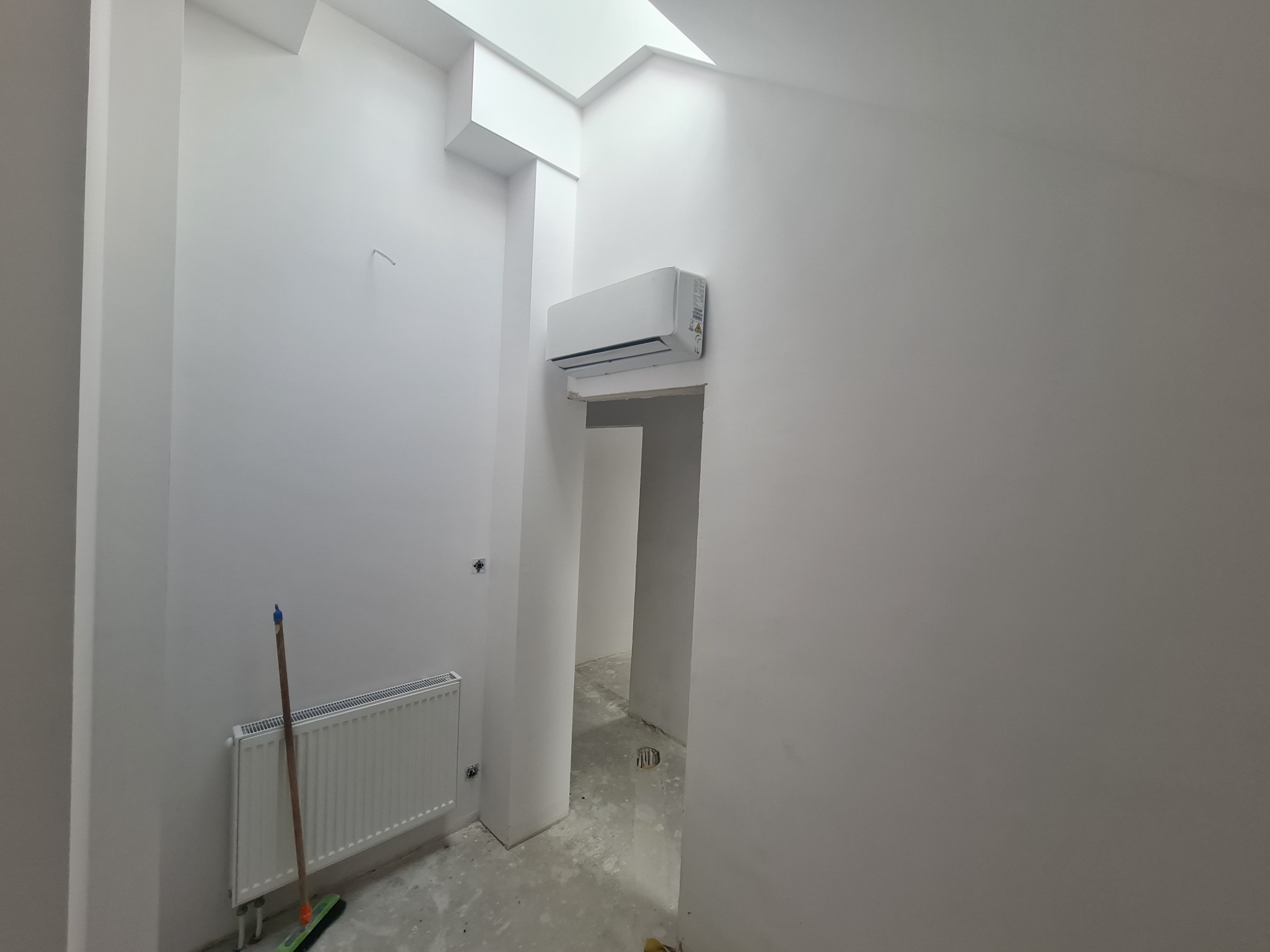 Biały klimatyzator ścienny zamontowany nad przejściem w surowym wnętrzu z grzejnikiem i świetlikiem, widoczne ślady po pracach wykończeniowych.