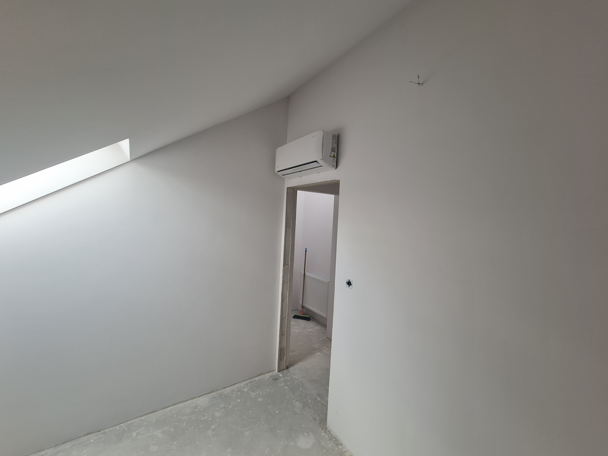 Wnętrze w trakcie wykańczania z zamontowanym klimatyzatorem na ścianie nad ościeżnicą. Widoczny fragment okna dachowego, szpachlowane ściany i podłoga. W przejściu widoczny grzejnik i miotła.