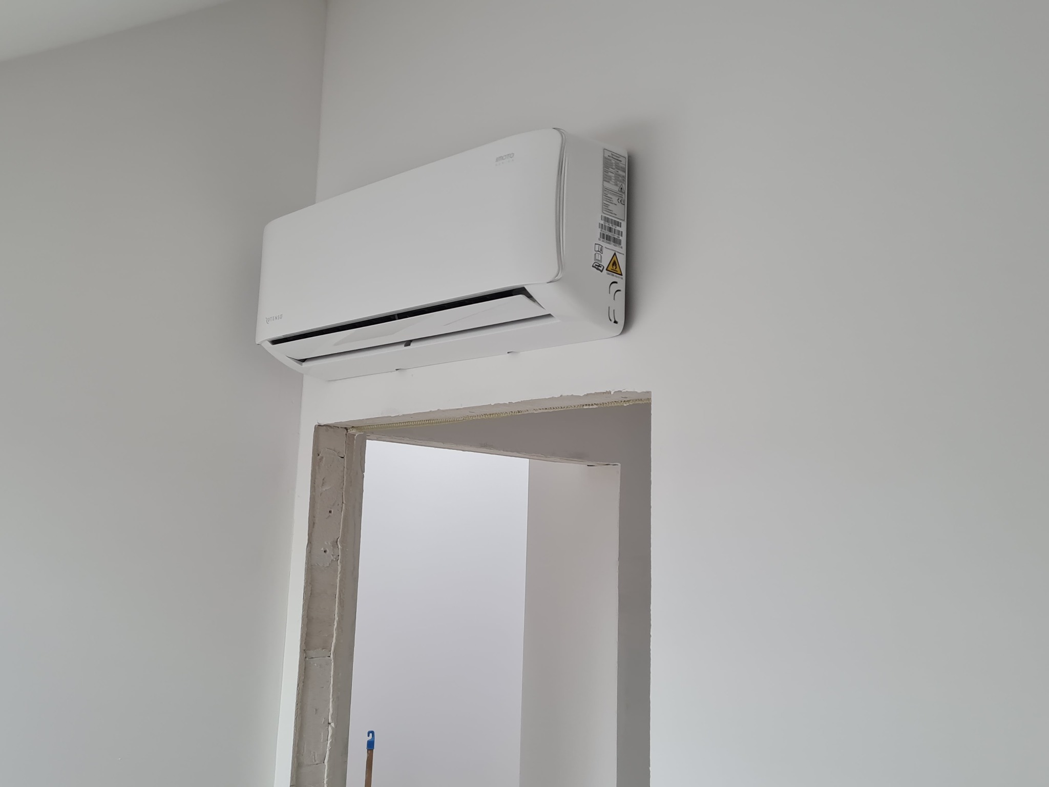 Biały klimatyzator zamontowany na białej ścianie nad surowym otworem drzwiowym, widoczne naklejki informacyjne na boku urządzenia.