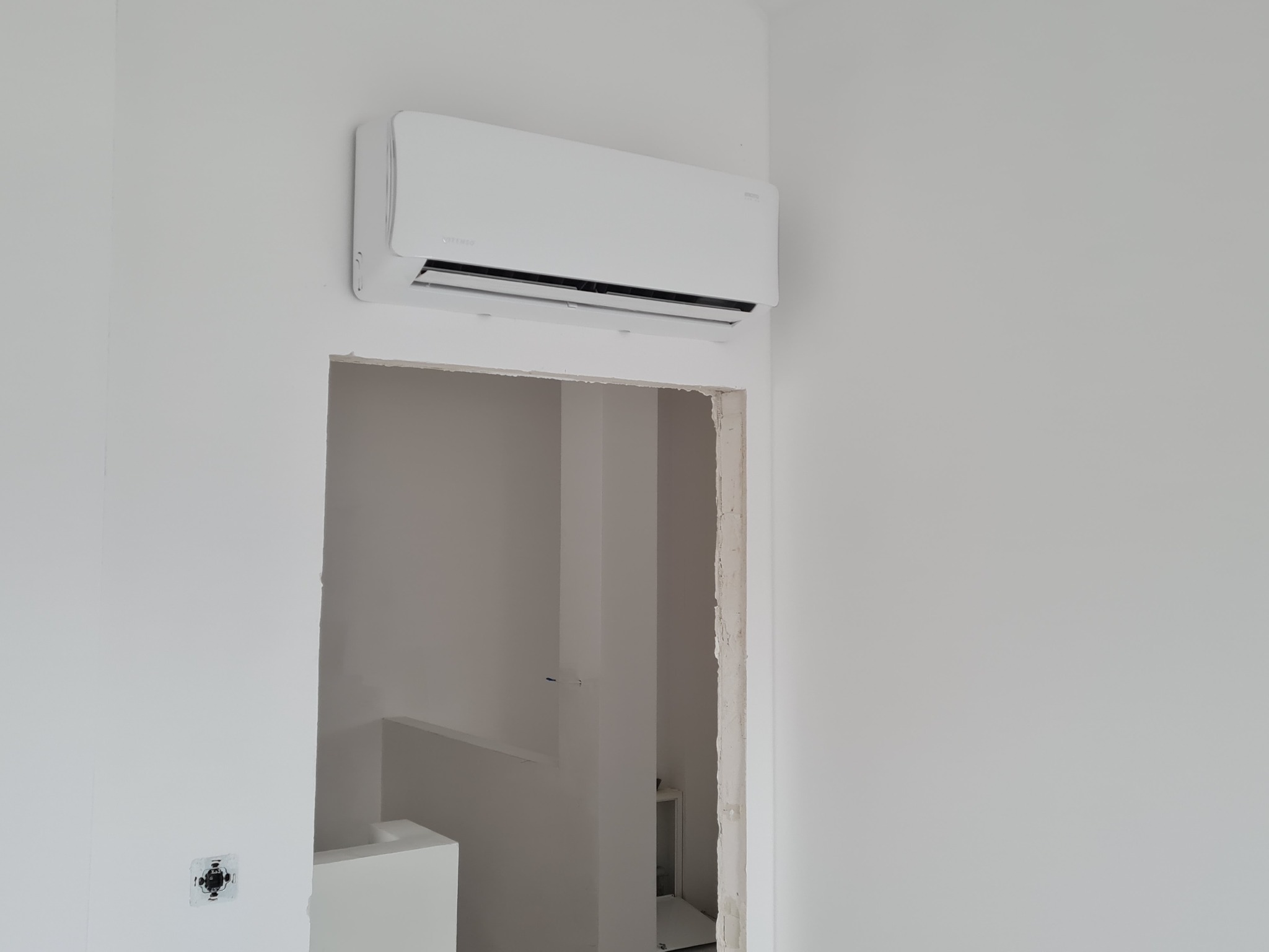 Biały klimatyzator Vitemo zamontowany na ścianie w nowym, jeszcze niewykończonym pomieszczeniu z widocznym otworem drzwiowym i puszką elektryczną.