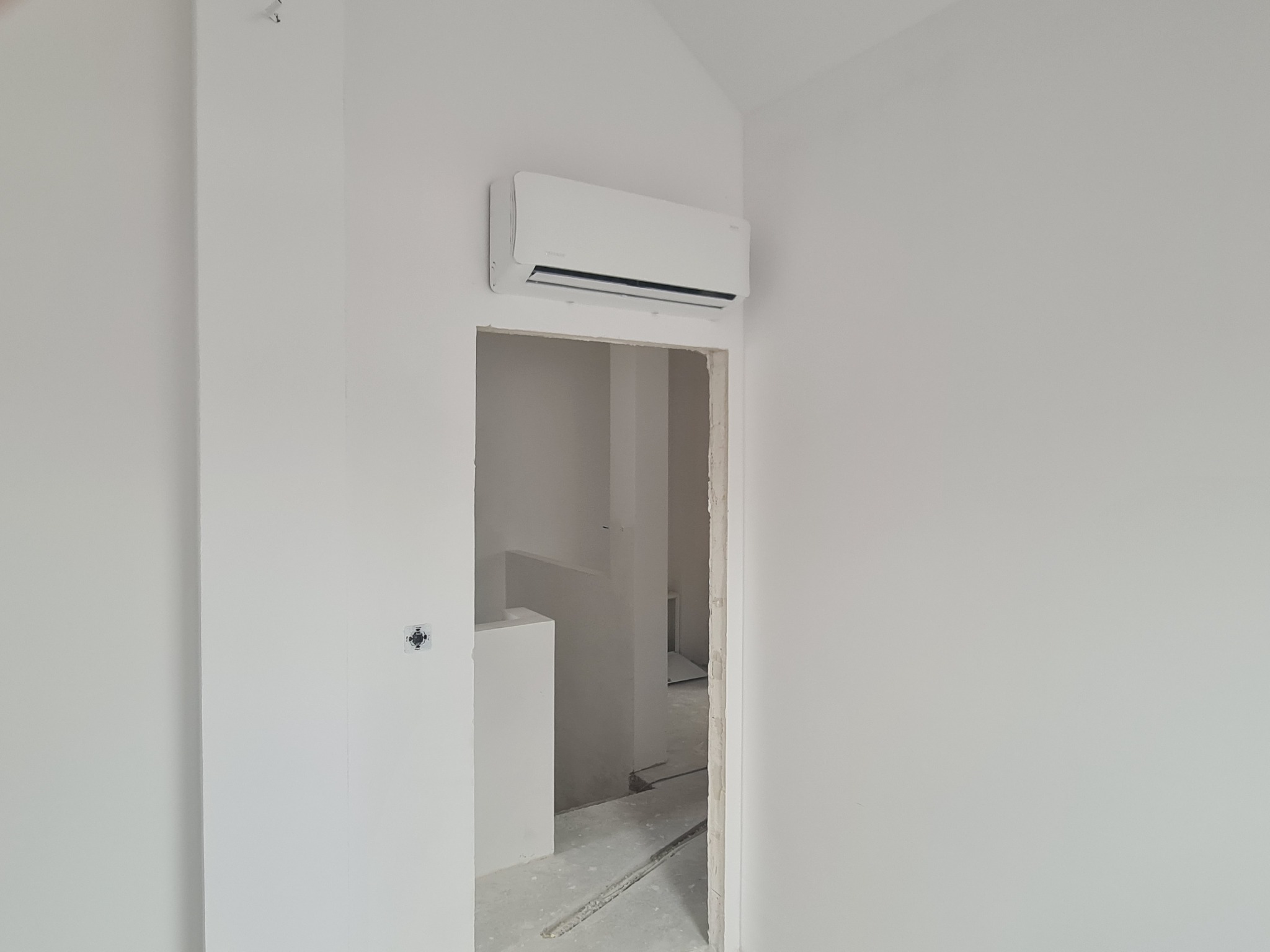 Biała jednostka klimatyzacyjna zamontowana nad ościeżnicą drzwiową w surowym, białym pomieszczeniu w trakcie remontu.