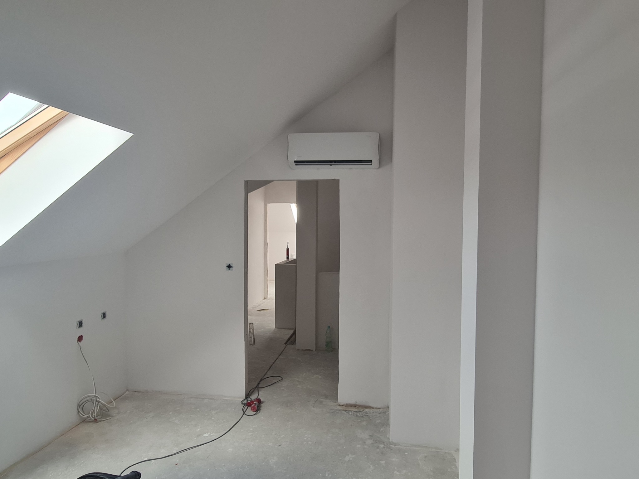 Biały klimatyzator ścienny zamontowany w jasnym, surowym wnętrzu poddasza ze skośnym oknem dachowym, widoczne kable i otwory w ścianie sugerujące prace instalacyjne.
