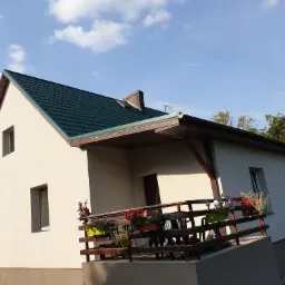 Jasny dom jednorodzinny z zielonym dachem, wykończoną elewacją i drewnianym balkonem ozdobionym kolorowymi kwiatami w doniczkach, widoczny fragment ogrodu i błękitne niebo.