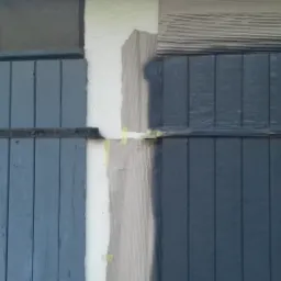 Fragment odnowionych, pomalowanych na szaro-niebiesko, drewnianych wrót garażowych z metalowymi zawiasami, zabezpieczonych taśmą malarską przy krawędzi styku z białą ścianą budynku.