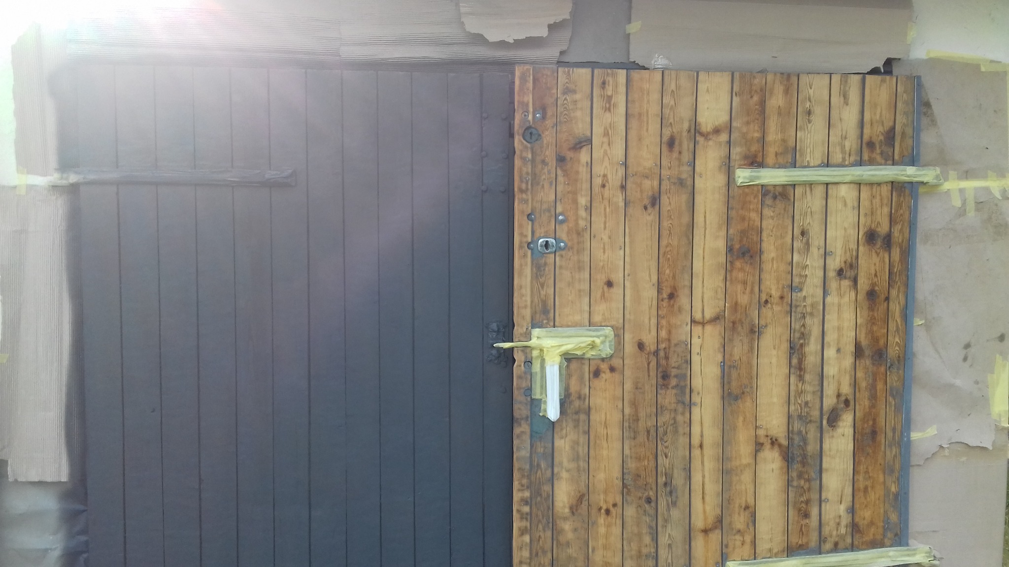 Drewniane wrota garażowe, połowa pomalowana na szaro, druga w naturalnym kolorze drewna, z widocznymi zabezpieczeniami z taśmy malarskiej.