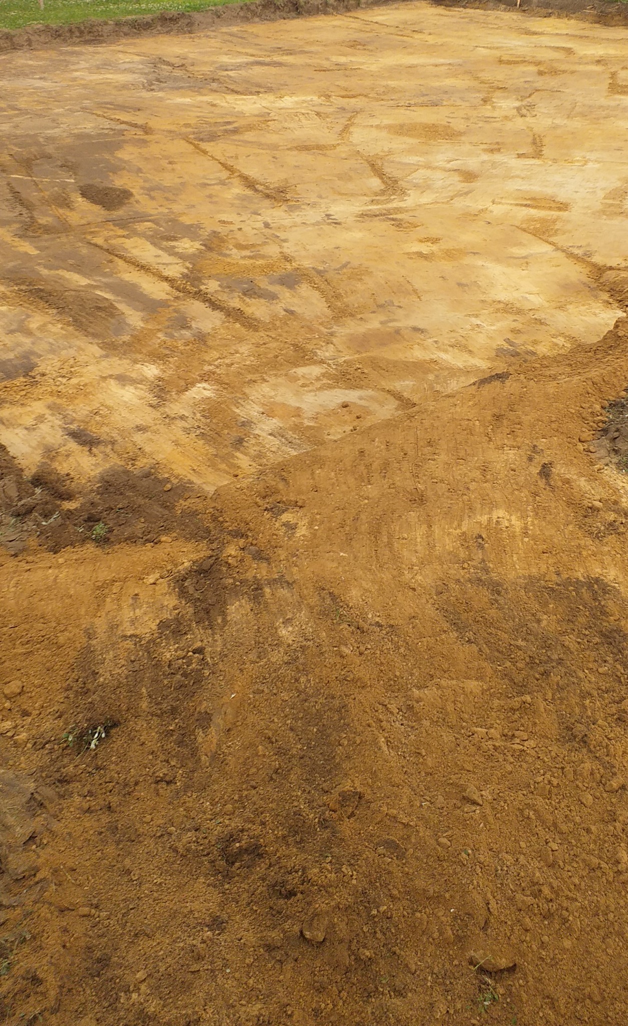 Przygotowany teren pod budowę: wyrównana, piaszczysta ziemia z widocznymi śladami po maszynach budowlanych, w tle zielony pas trawy.