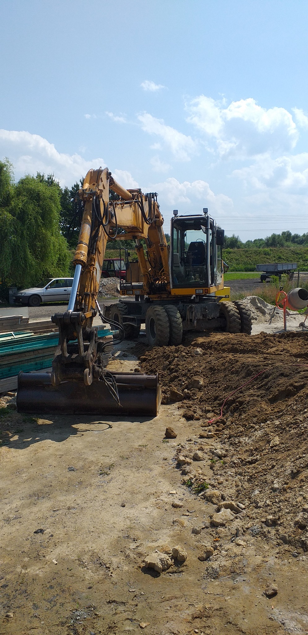 Żółta koparka kołowa z łyżką na placu budowy, widoczna świeżo naruszona ziemia, w tle drzewa i błękitne niebo z chmurami.
