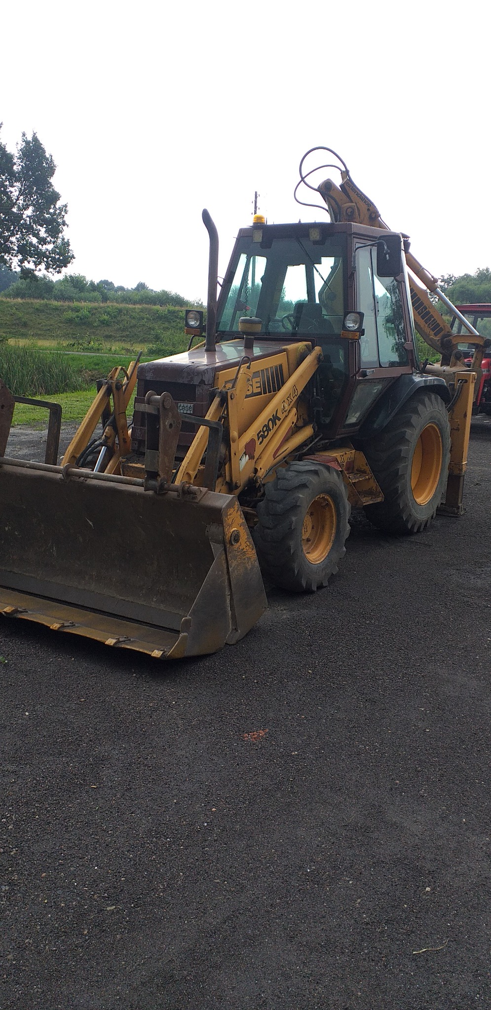 Żółto-czarna koparko-ładowarka Case 580K 4x4 zaparkowana na czarnym asfalcie, z widocznym łyżką z przodu i ramieniem koparki z tyłu, na tle zieleni.