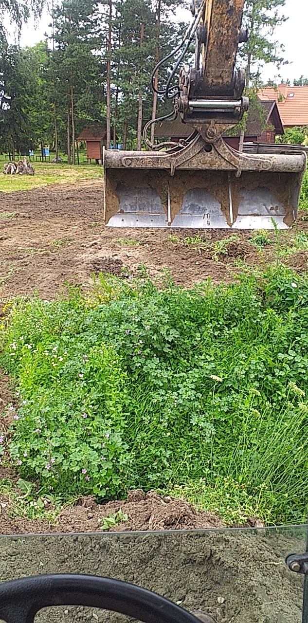 Łyżka koparki nad kępą zieleni, przygotowany teren pod wykop z widocznymi zabudowaniami w tle.
