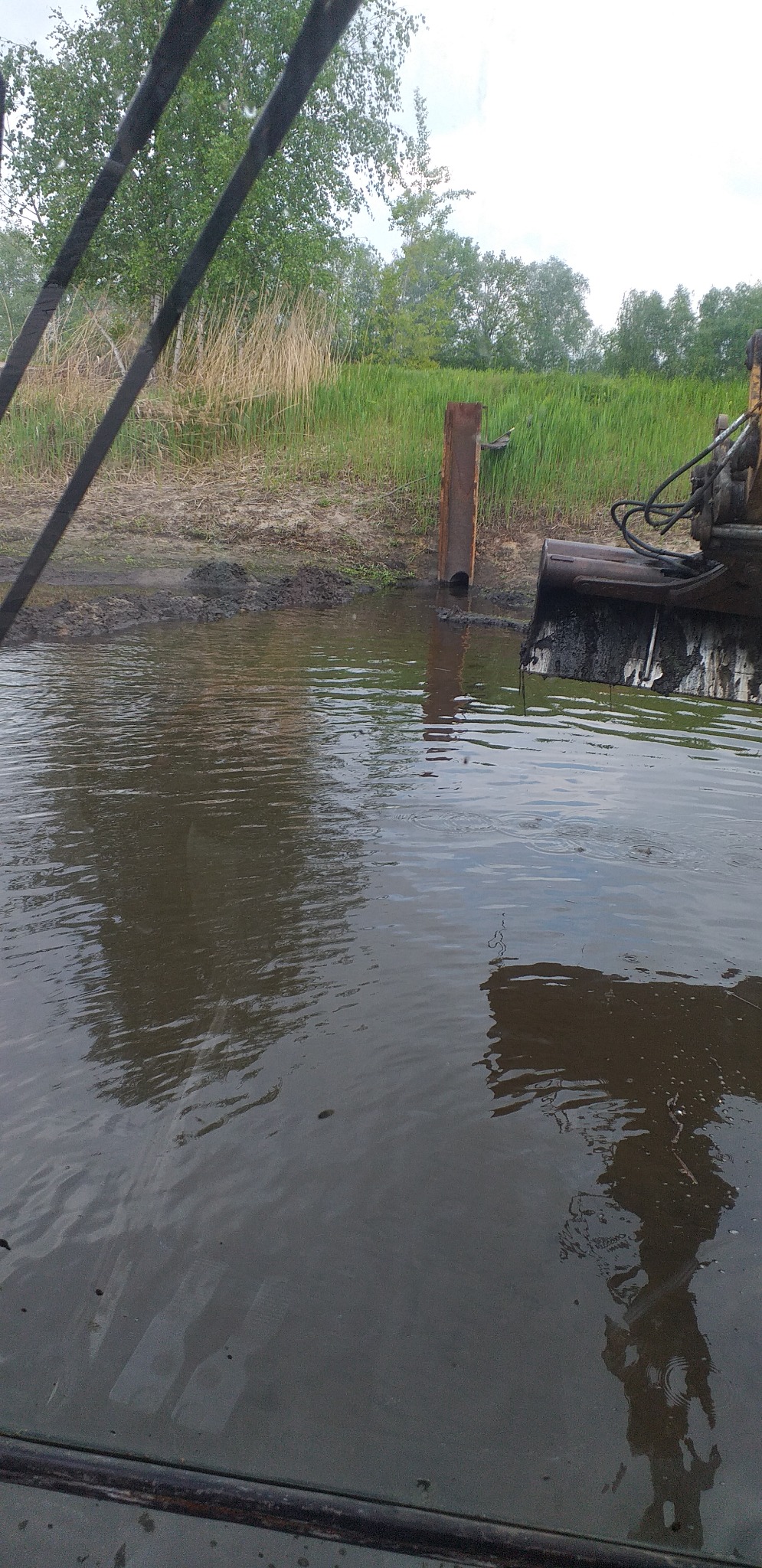 Koparka zanurzona w wodzie, prawdopodobnie pogłębia dno zbiornika, obok wbita stalowa rura, brzeg porośnięty trawą i trzciną.