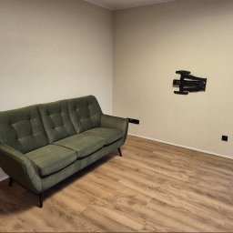 Dominik Kulej DomKul - Wnętrze pokoju po odświeżeniu, z zieloną sofą na parkiecie, uchwytem do telewizora na ścianie i widokiem na balkon przez okno.