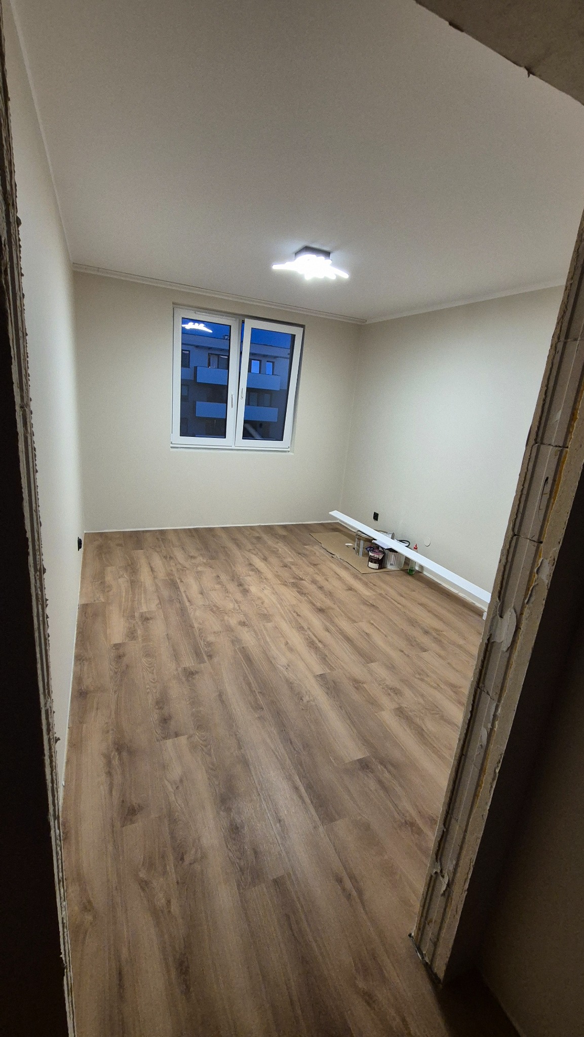 Pusty pokój z panelami podłogowymi w odcieniach brązu, kremowe ściany, białe okno z widokiem na budynek, listwy przypodłogowe, widoczny fragment ościeżnicy w trakcie prac.