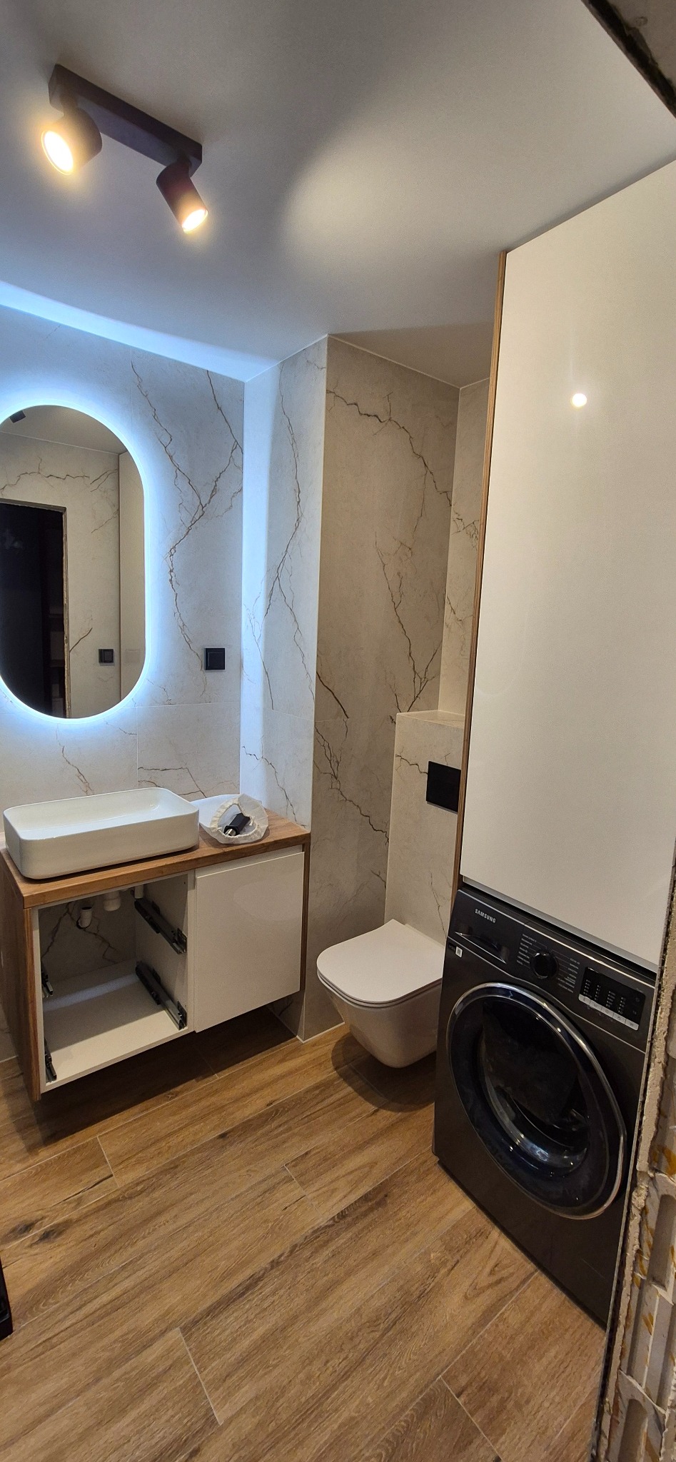 Nowoczesna łazienka z umywalką nablatową, podświetlanym lustrem, toaletą wiszącą i pralką w zabudowie, wykończona płytkami imitującymi marmur i drewno.