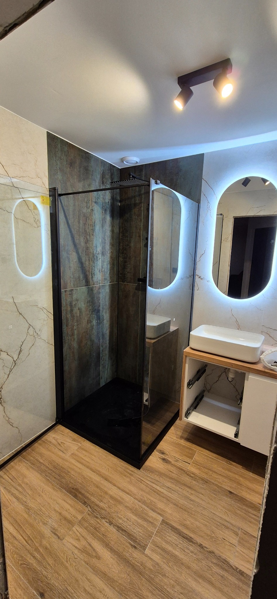 Nowoczesna łazienka z kabiną prysznicową typu walk-in z czarnymi profilami, podświetlanymi lustrami owalnymi i drewnopodobną podłogą ułożoną w jodełkę.