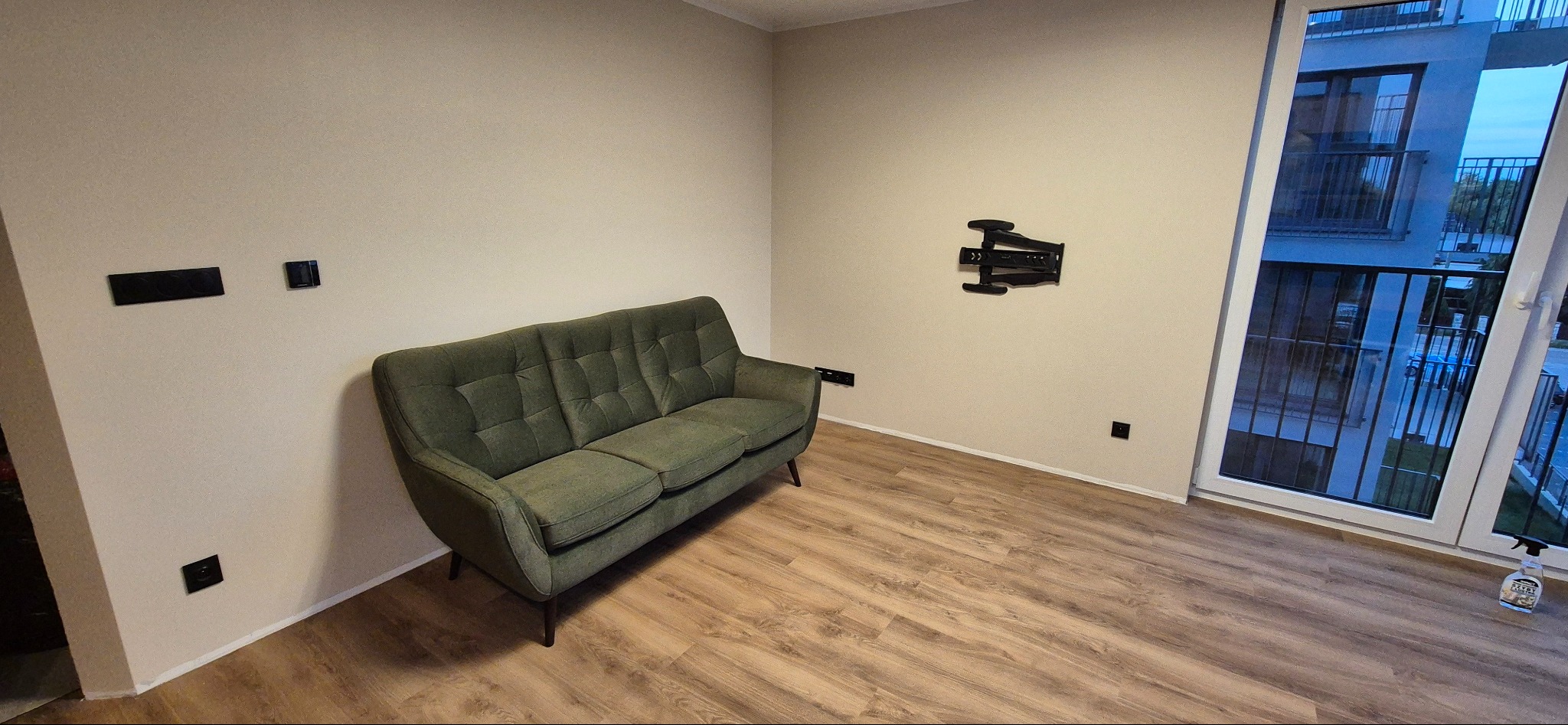 Wnętrze pokoju po odświeżeniu, z zieloną sofą na parkiecie, uchwytem do telewizora na ścianie i widokiem na balkon przez okno.
