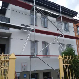 Ocieplona elewacja budynku w trakcie malowania, widoczne rusztowania, okna zabezpieczone folią, żółte ogrodzenie w pierwszym planie.