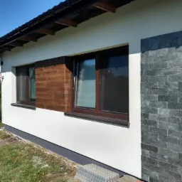 Wykończona elewacja domu jednorodzinnego z fragmentami w kolorze białym, drewnopodobnym i z szarej kamiennej okładziny. Widoczna instalacja antenowa na dachu sąsiedniego budynku.