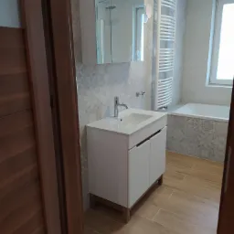 Nowoczesna łazienka z białymi meblami łazienkowymi, grzejnikiem drabinkowym i płytkami w geometryczne wzory, widok zza otwartych, drewnianych drzwi.