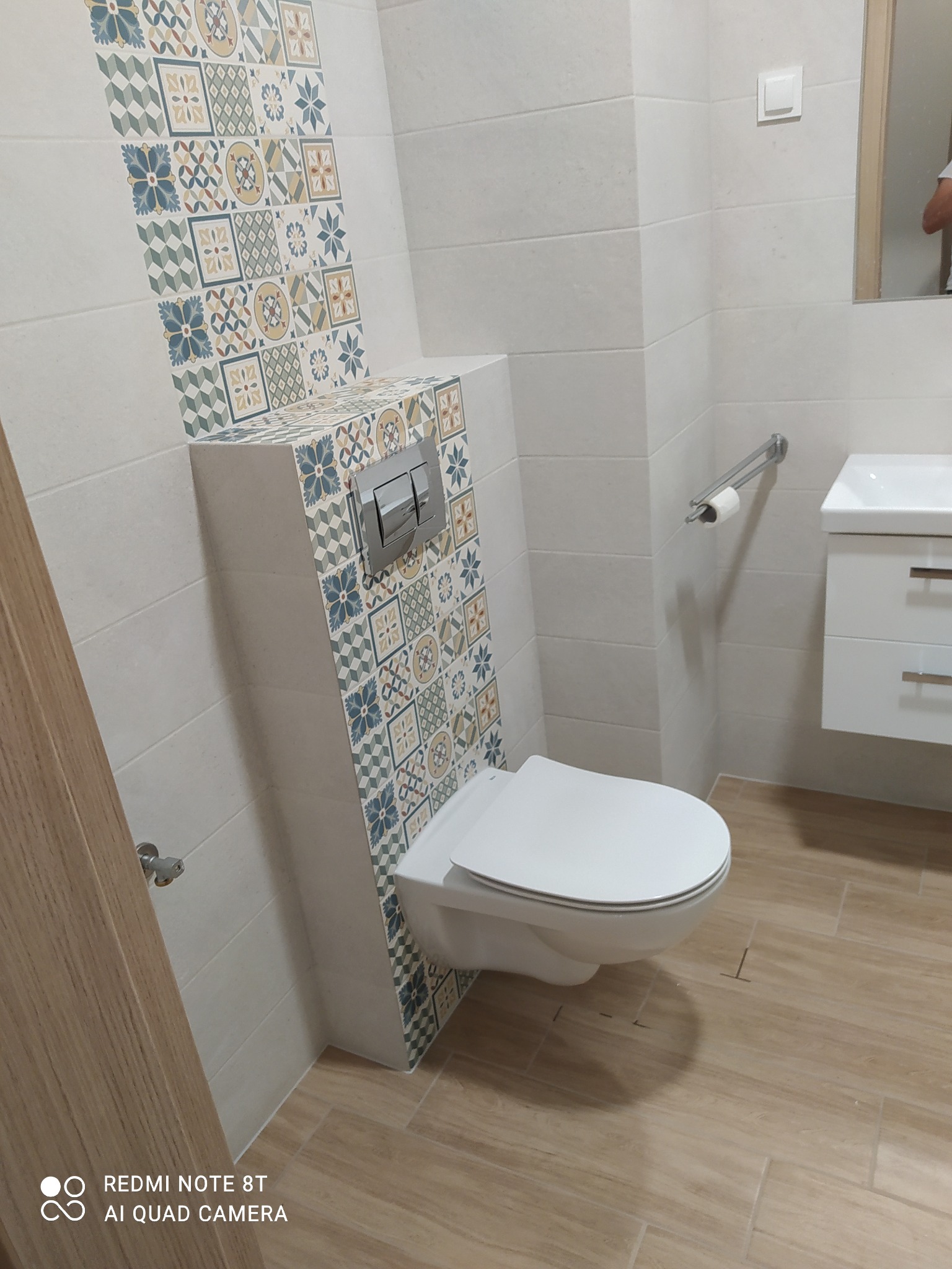 Nowoczesna łazienka z podwieszaną toaletą i zabudową stelaża w płytkach dekoracyjnych, jasne płytki na ścianach i podłodze imitujące drewno.