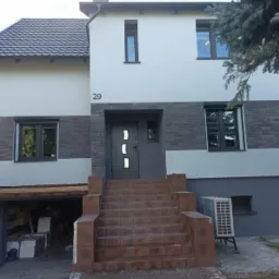 Dwupiętrowy dom w Bydgoszczy po remoncie elewacji, z szarą cegłą klinkierową na dolnej części fasady i jasnoszarym tynkiem na g&oacute;rze, z numerem 29 nad wejściem i schodami z brązowej cegły.