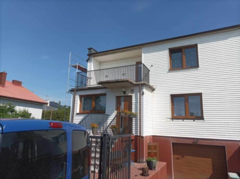 Dom jednorodzinny z elewacją z białego sidingu, brązowymi oknami i balkonem z czarną balustradą, częściowo zasłonięty przez niebieskiego vana, z widocznym rusztowaniem z boku.