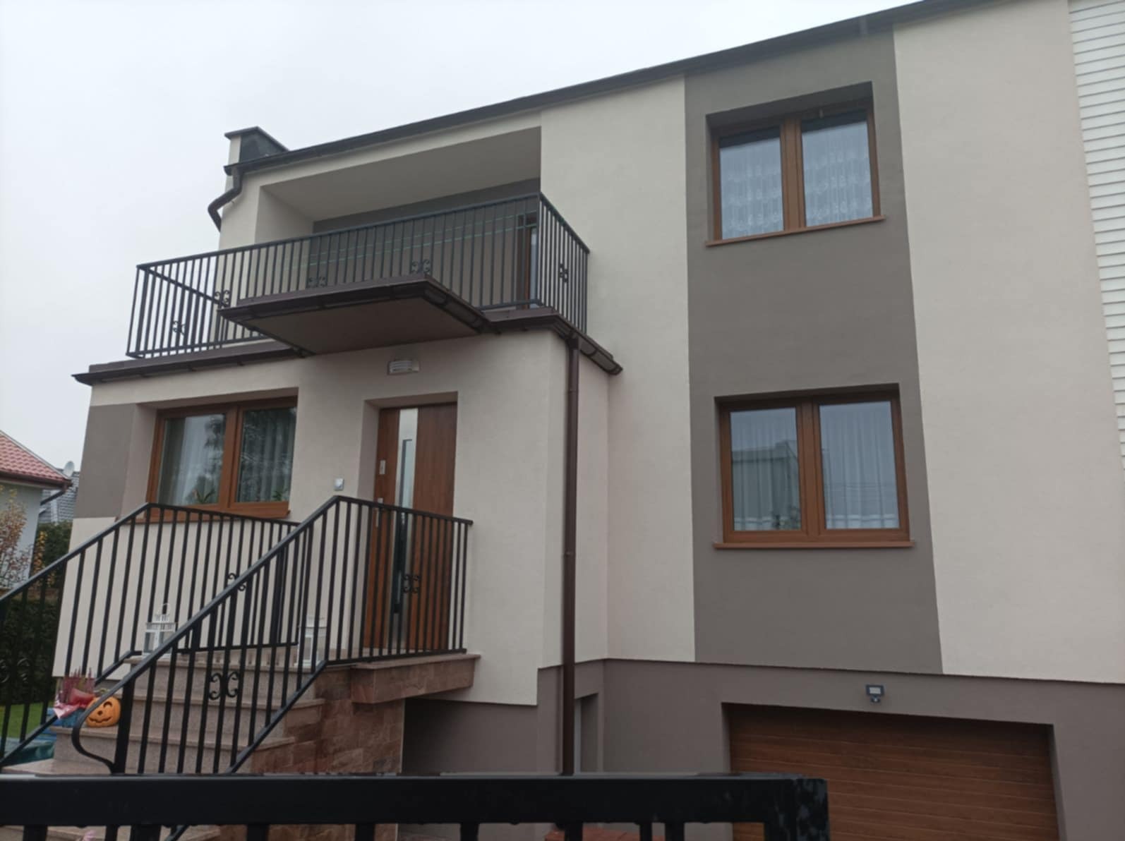 Dwukondygnacyjny dom z balkonem, elewacja w odcieniach beżu i szarości, brązowe okna, schody wejściowe z czarną balustradą, widok z dołu.