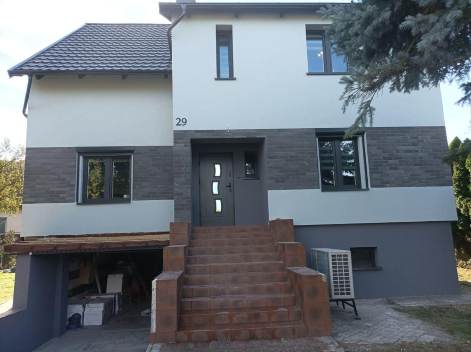 Dwupiętrowy dom w Bydgoszczy po remoncie elewacji, z szarą cegłą klinkierową na dolnej części fasady i jasnoszarym tynkiem na górze, z numerem 29 nad wejściem i schodami z brązowej cegły.
