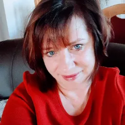 Portret kobiety w czerwonej bluzce z krótkimi brązowymi włosami i niebieskimi oczami, siedzącej na ciemnej skórzanej kanapie.