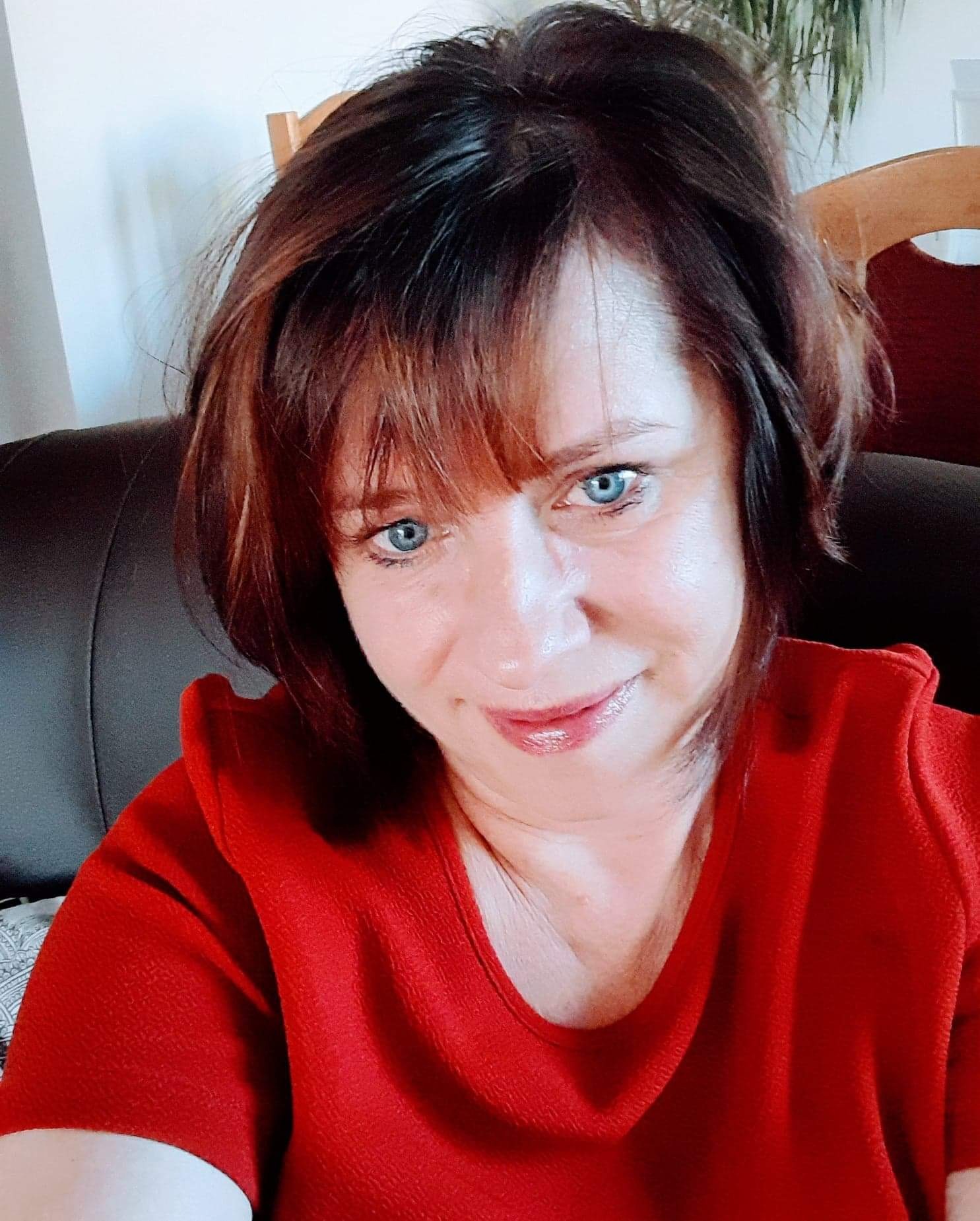 Portret kobiety w czerwonej bluzce z krótkimi brązowymi włosami i niebieskimi oczami, siedzącej na ciemnej skórzanej kanapie.