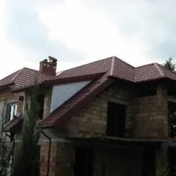 Dom w trakcie budowy z częściowo wykonanym dachem z czerwonej blachodachówki, widoczny pracownik na kominie i niezakończone elewacje.