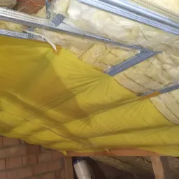 Ocieplenie poddasza wełną mineralną, zabezpieczone żółtą folią paroizolacyjną i zamocowane do metalowego rusztu, widoczne cegły ściany bocznej.