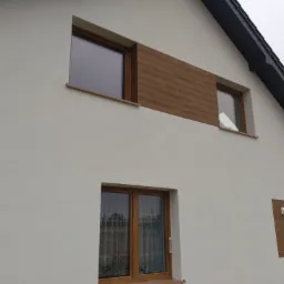 Biała elewacja domu jednorodzinnego z drewnopodobnymi panelami dekoracyjnymi wokół okien, widok z dołu na fragment ściany szczytowej z oknami i fragmentem dachu pokrytego czarną dachówką.