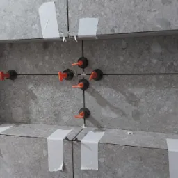 Półka wnękowa w trakcie układania płytek w szarym odcieniu, z widocznymi krzyżykami dystansowymi, pomarańczowymi klipsami poziomującymi i taśmą maskującą.