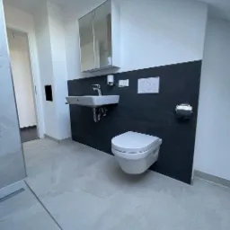 Nowoczesna łazienka z szarą płytką podłogową, wiszącą miską WC, umywalką i szafką z lustrem na czarnej ścianie akcentującej, widoczne przewody elektryczne.