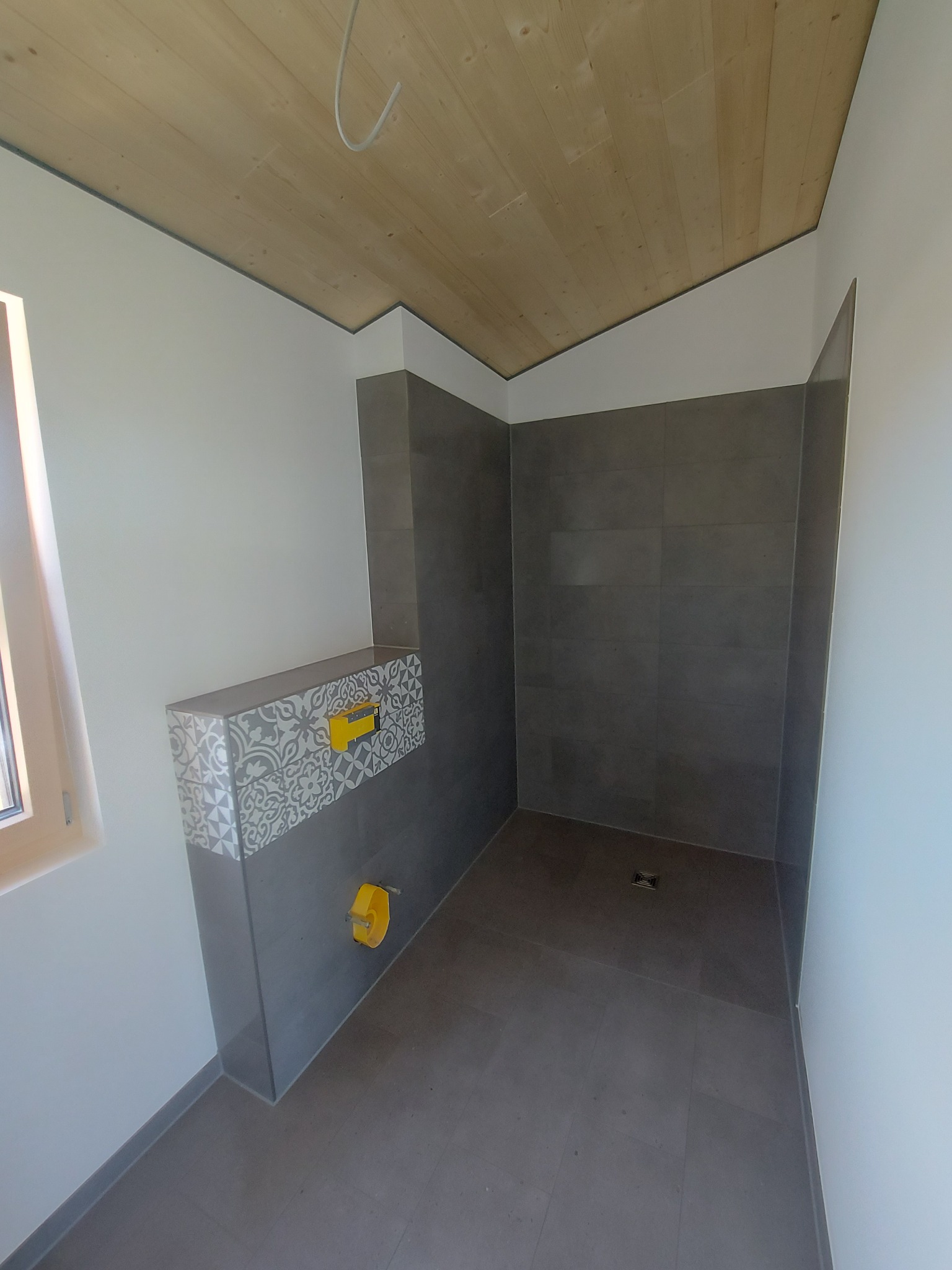 Nowoczesna kabina prysznicowa z szarymi płytkami i drewnianym sufitem, widoczny ozdobny panel z mozaiką i żółtym elementem instalacji.