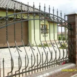 Kute ogrodzenie z ceglanymi słupkami i dekoracyjnymi elementami. Falista linia górna. Tło: dom z garażem i okno z ozdobną kratką. Ujęcie z bliska.