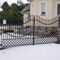 Czarna, kuta brama wjazdowa z dekoracyjnymi elementami, osadzona na kamiennych słupkach. W tle ośnieżony teren i jasny budynek z oknami.