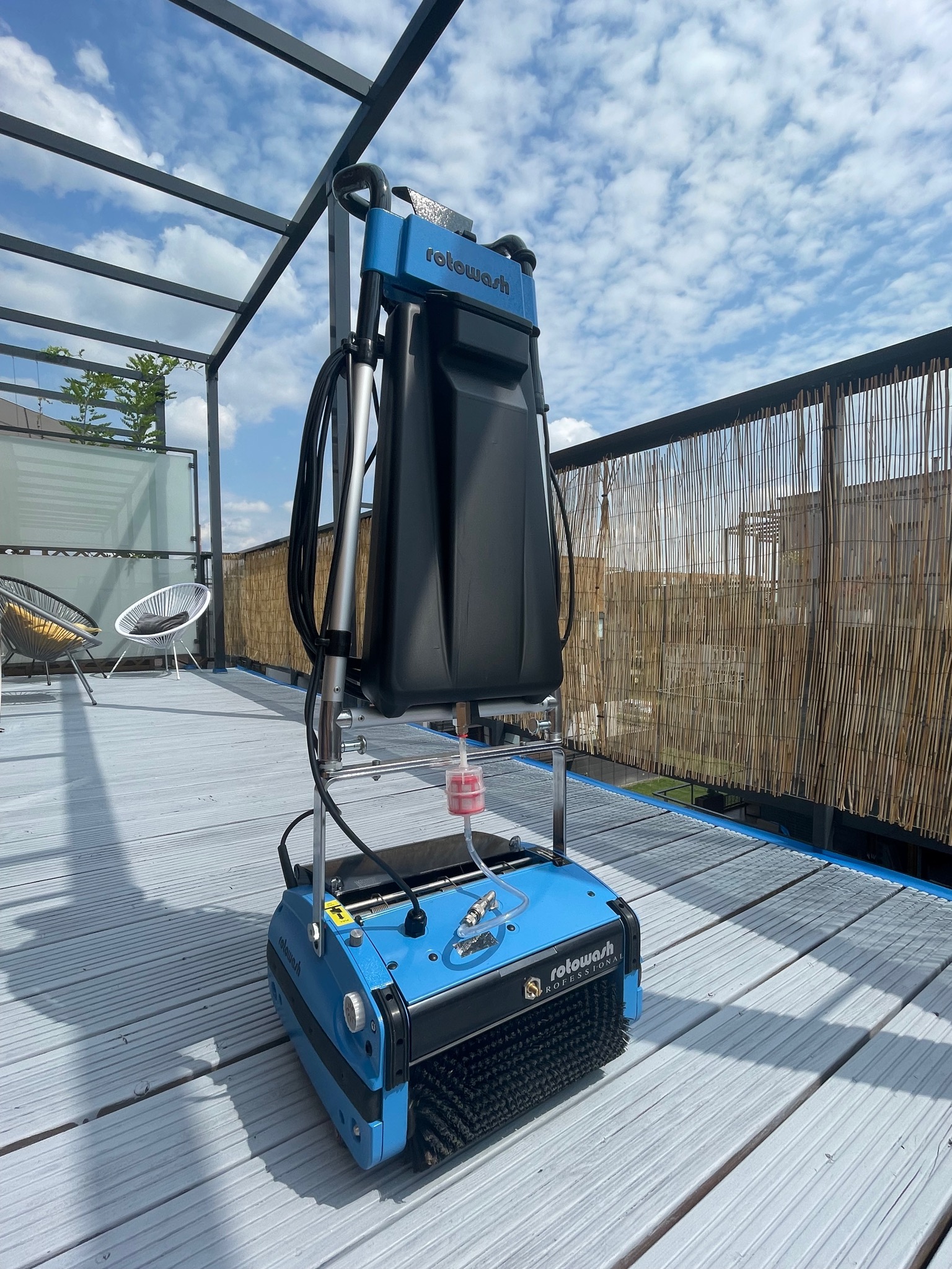 Profesjonalna maszyna Rotowash do czyszczenia powierzchni na szarym tarasie z drewnianą podłogą, widok z niskiej perspektywy, błękitne niebo w tle.