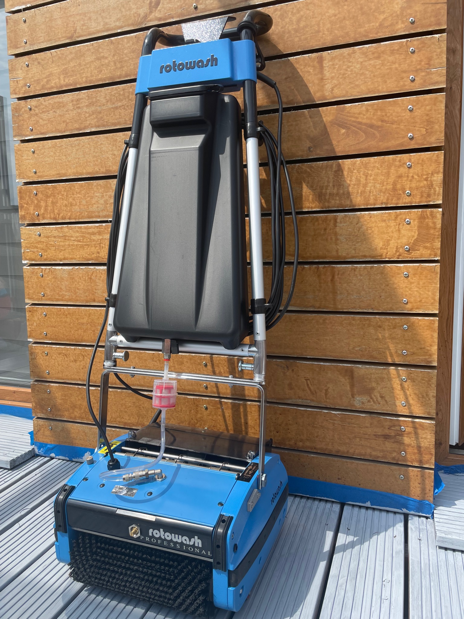 Niebieska maszyna czyszcząca Rotowash Professional stojąca na szarej podłodze tarasu, z drewnianą ścianą w tle, widoczny zbiornik na wodę i szczotki czyszczące.