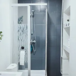 Nowoczesna łazienka z kabiną prysznicową, szarymi płytkami i białymi elementami wyposażenia, widok na umywalkę i toaletę.