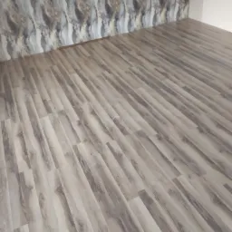 Ułożone panele podłogowe w odcieniach szarości i beżu, widoczne na tle wzorzystej tapety w podobnej kolorystyce, z fragmentem białej ściany w rogu pomieszczenia.