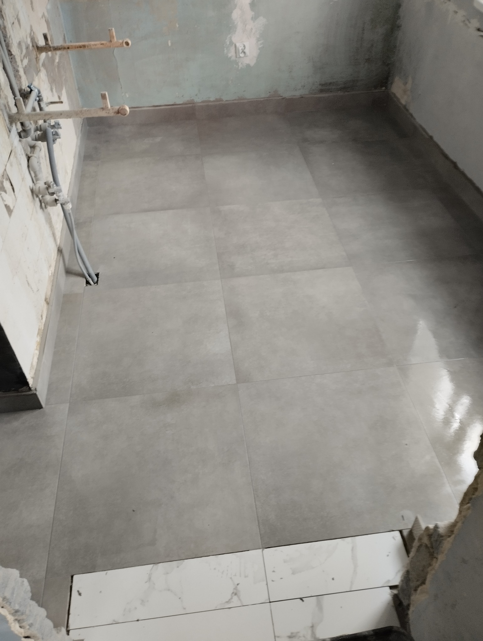 Wyłożona szarymi płytkami podłoga w trakcie remontu kuchni, widoczne rury instalacji wodnej przy ścianie oraz fragment niezakończonej podłogi z białymi płytkami imitującymi marmur.